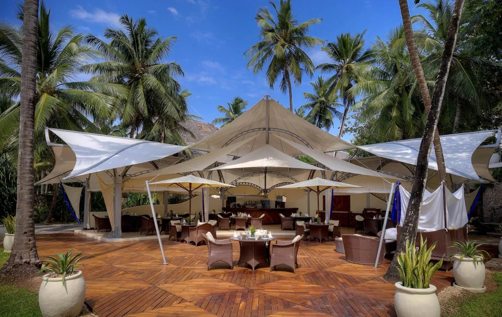 Open-air beachfront restaurant with white canopy, wooden deck seating, and palm trees at The Sails Beach Bar & Restaurant in Diani Beach.