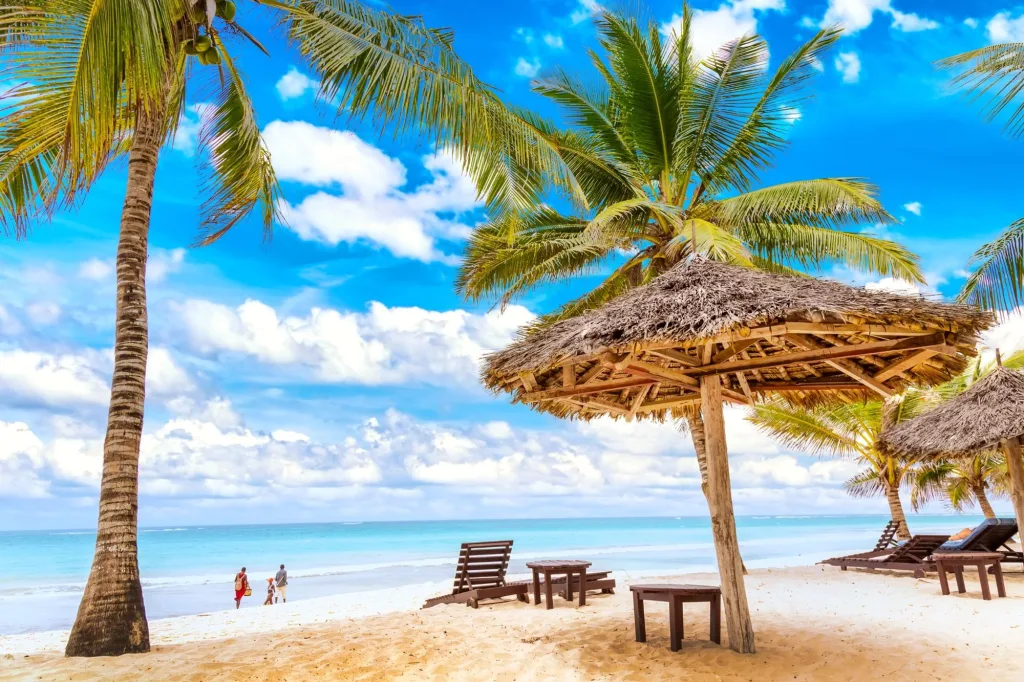 Diani beach scene with palm trees, thatch umbrellas, and people walking on the sand. Best Beach Spots in Diani.