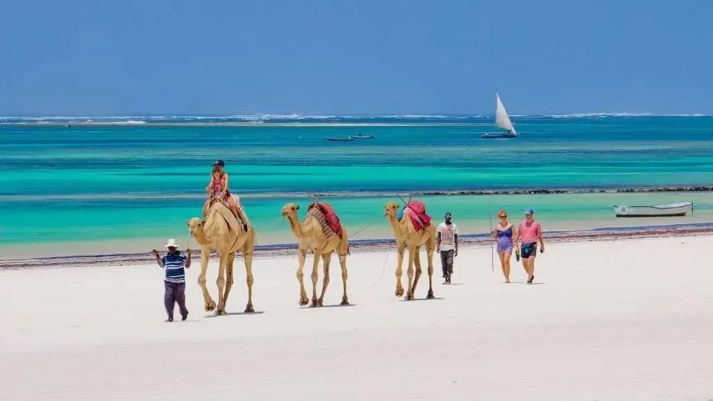Diani beach scene with palm trees, thatch umbrellas, and people walking on the sand. Best Beach Spots in Diani.
