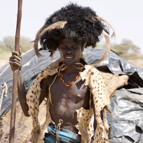 African man wearing traditional animal skin clothing and headgear holding a spear in a rural setting